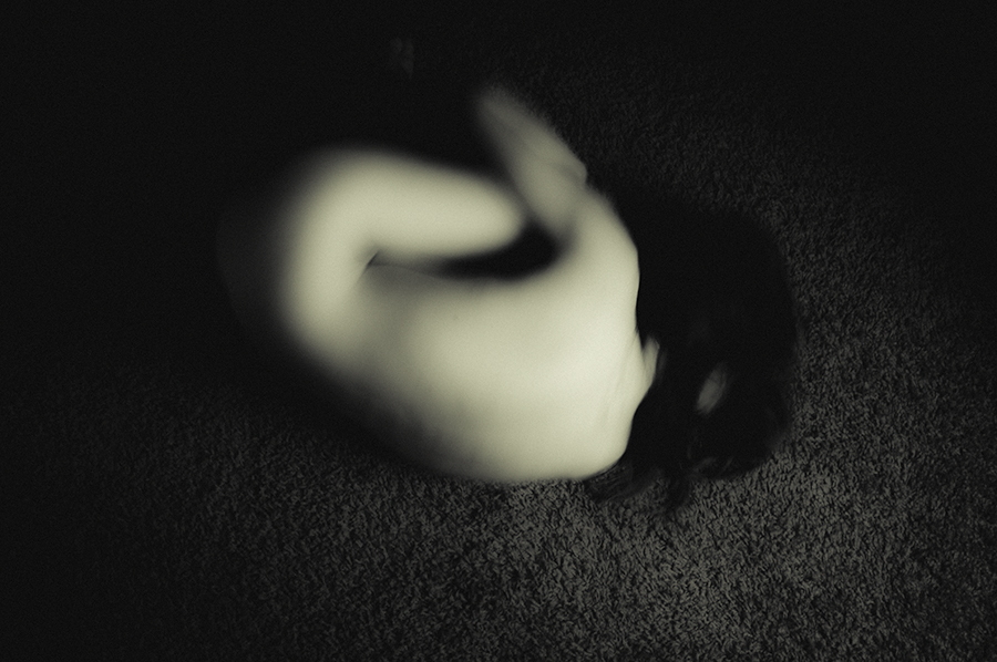 Blurred pale figure curled against a dark textured floor in a black-and-white photograph.