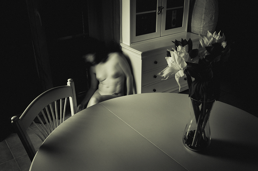 Blurred nude figure seated beside a round table and a vase of flowers in a dim room.