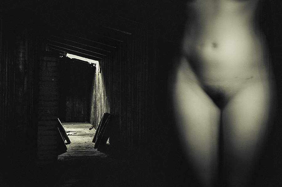 Blurred nude torso in the foreground of a dark room, with a doorway visible behind.