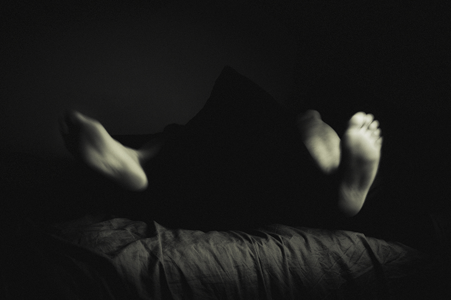 Two bare feet rise from a dark form above a rumpled bedspread.