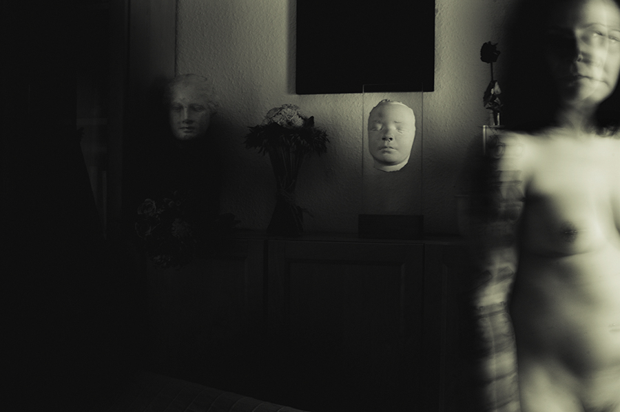 Blurred figure passing in front of a dresser with a pale mask in a dark room.