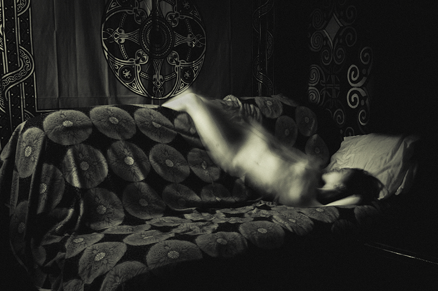 Blurred nude figure lying across a patterned sofa beneath wall hangings.