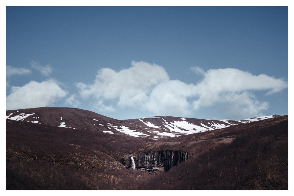 A waterfall drops between brown hills with patches of snow beneath white clouds - Pause by Juan Antonio Partal