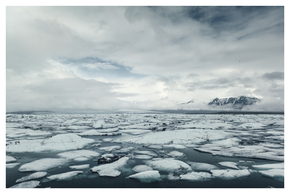 Broken ice floats on dark water with distant snowy mountains beneath low clouds - Pause by Juan Antonio Partal