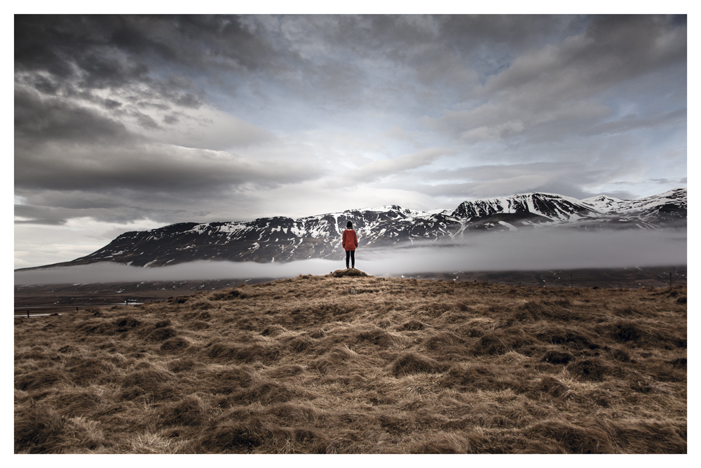 A person in a red jacket stands on a grassy rise facing snowy mountains and low clouds - Pause by Juan Antonio Partal