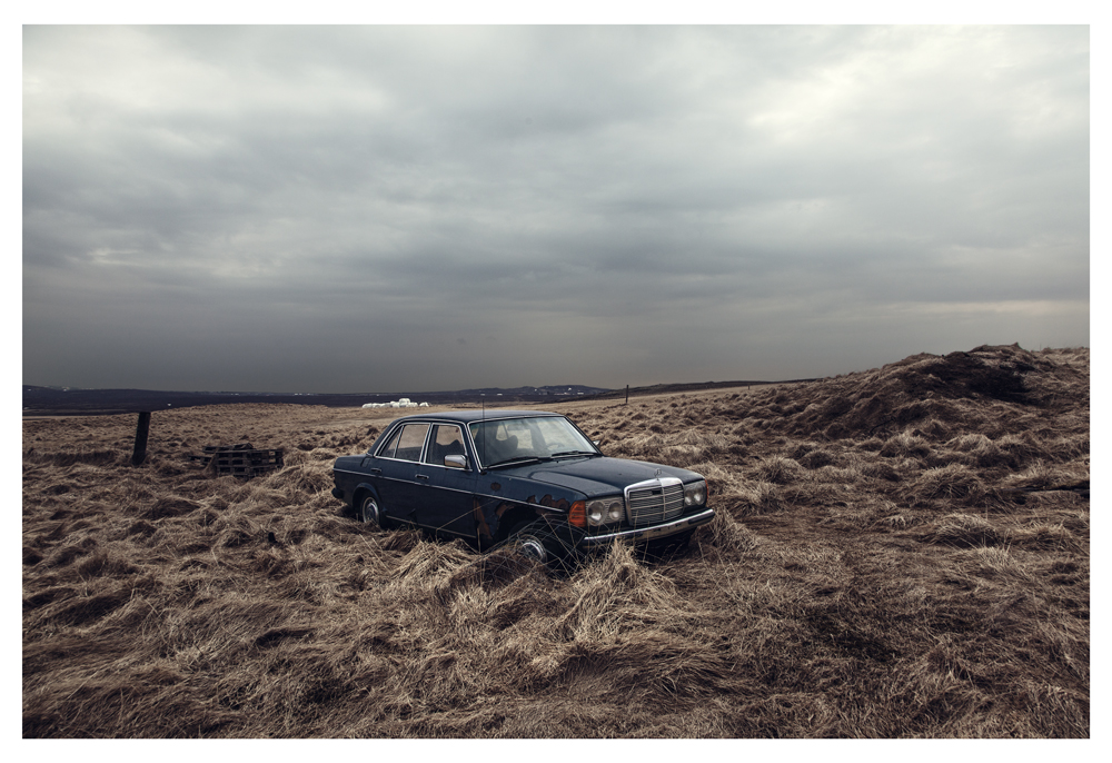 An old blue sedan sits in dry grass beneath a gray sky - Pause by Juan Antonio Partal