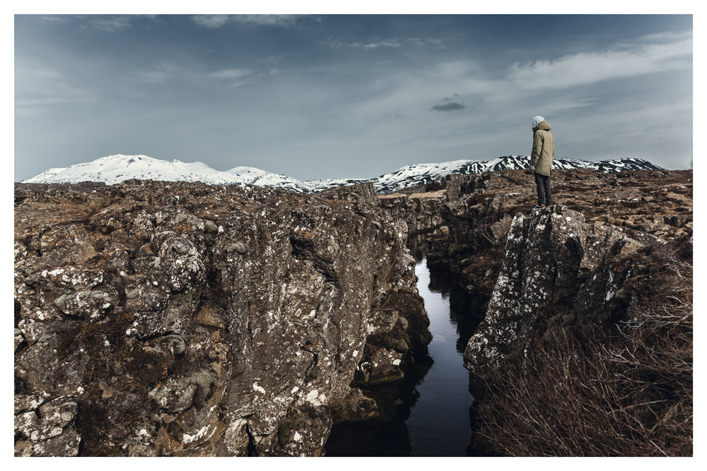 A person stands on rock beside a narrow water-filled fissure, with snowy hills beyond - Pause by Juan Antonio Partal