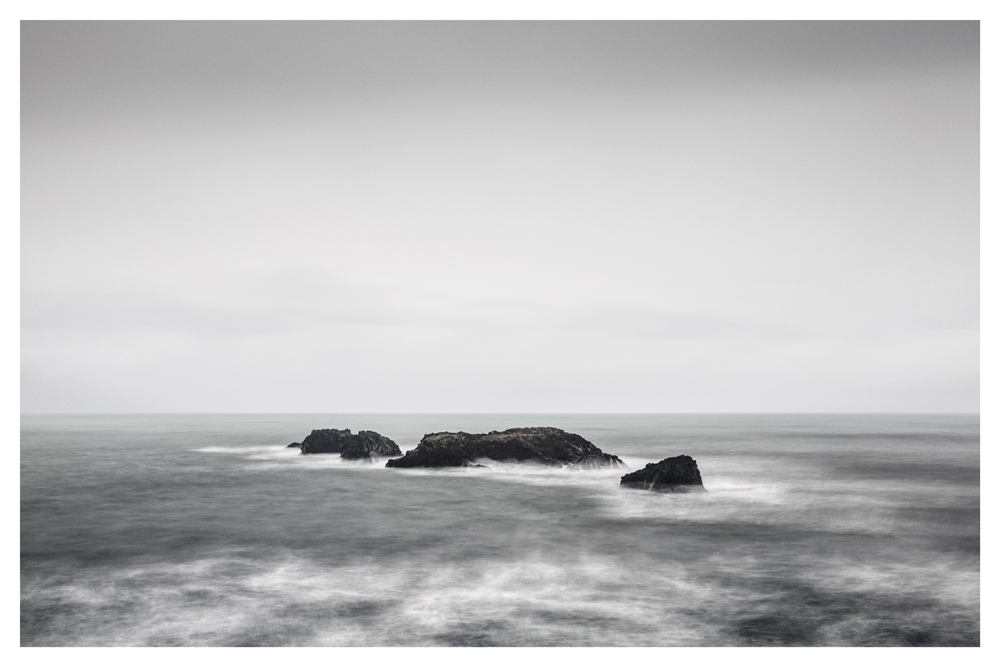 Dark rocks break the surface of a gray sea beneath a pale sky - Pause by Juan Antonio Partal