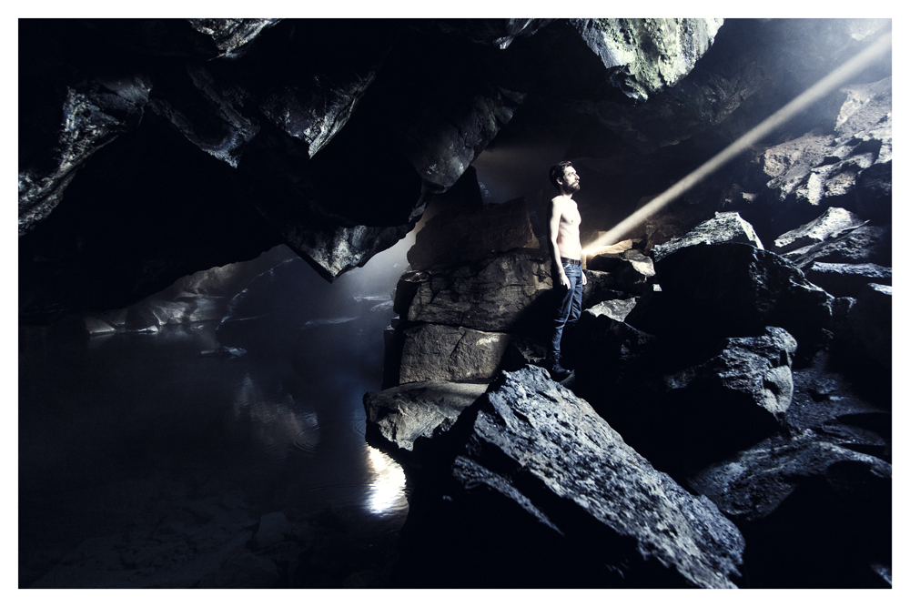 A shirtless man stands among rocks inside a dark cave, lit by a beam from above - Pause by Juan Antonio Partal