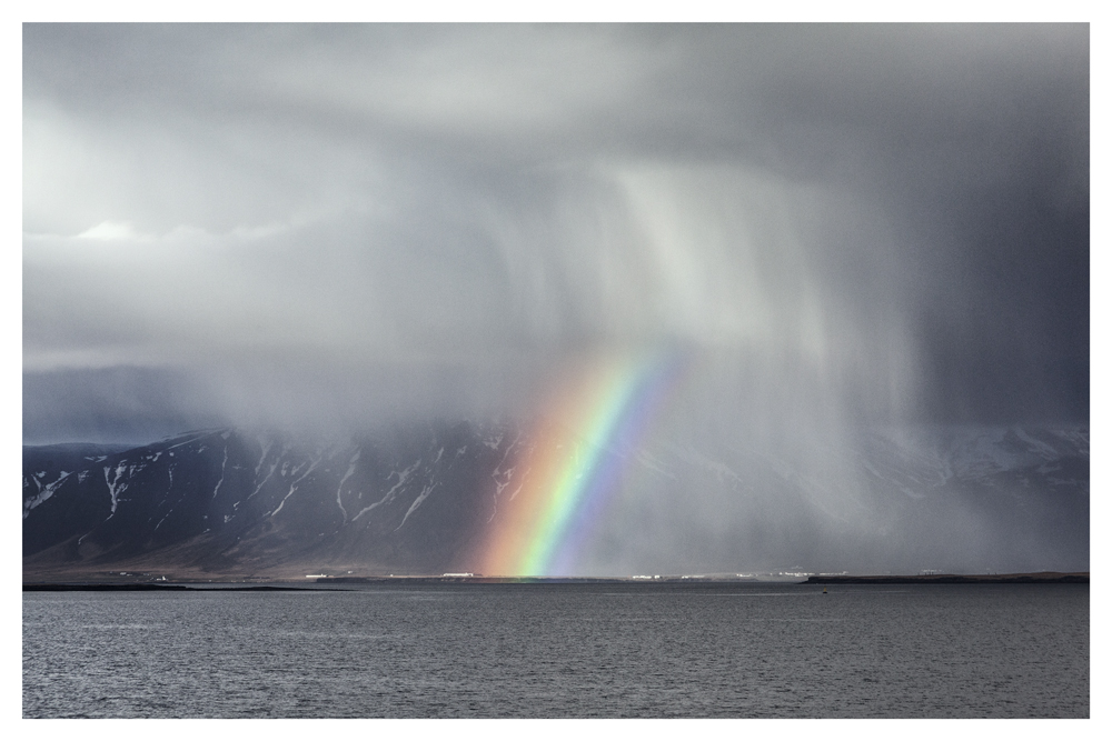 A rainbow descends beneath rain clouds over a mountain and a body of water - Pause by Juan Antonio Partal