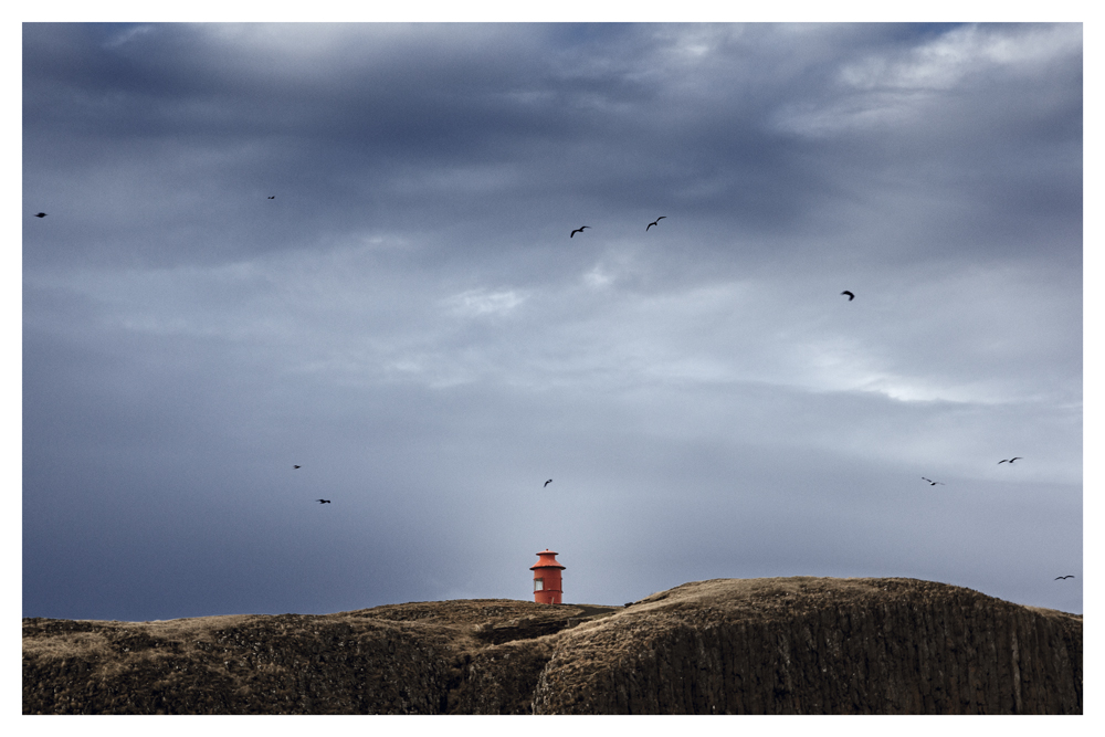 Birds circle above a small red lighthouse on a rocky hill beneath storm clouds - Pause by Juan Antonio Partal