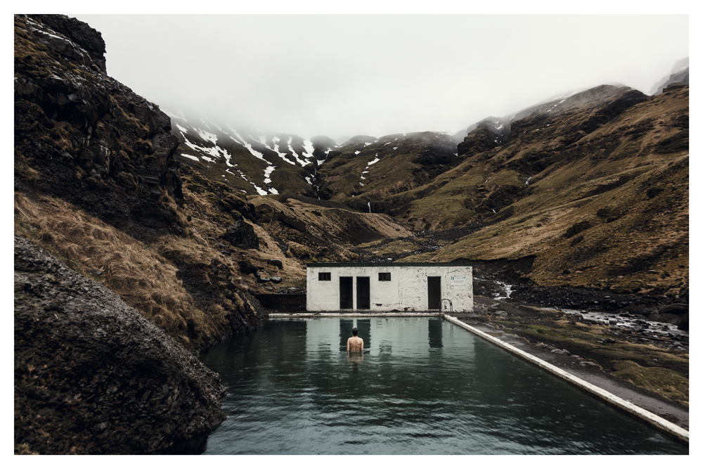 A person stands in an outdoor pool beside a white building, with snowy mountains behind - Pause by Juan Antonio Partal