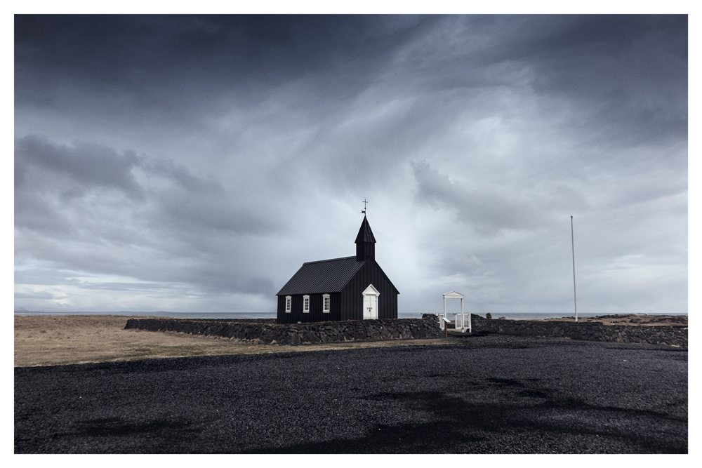 A small black church stands alone beneath a wide, cloud-filled sky - Pause by Juan Antonio Partal