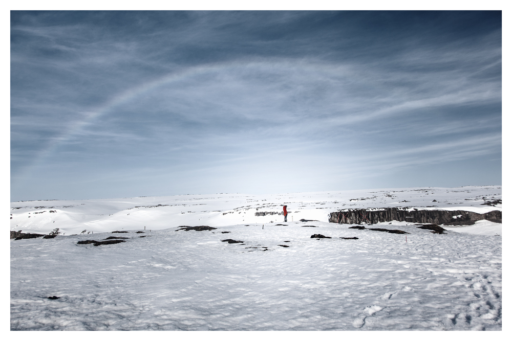 A person in a red jacket stands on a snowy plain beneath a faint rainbow in the sky - Pause by Juan Antonio Partal