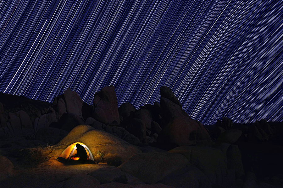 Star trails streak above a glowing tent and desert rock formations.