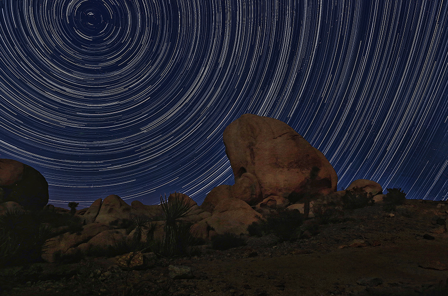 Circular star trails above a large boulder in a dark desert landscape.