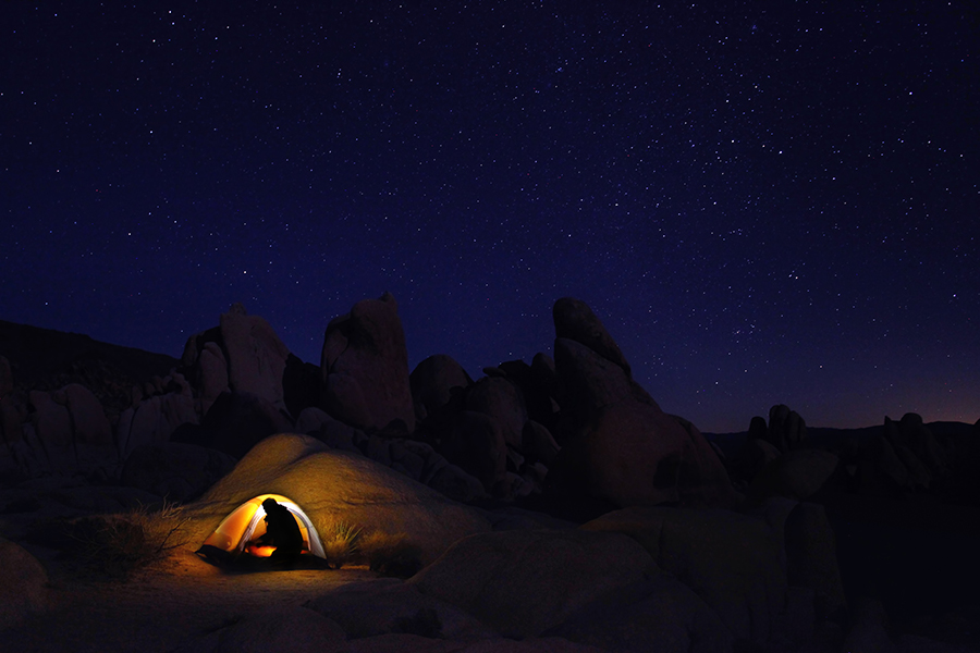 Glowing tent beneath stars and tall desert boulders at night.