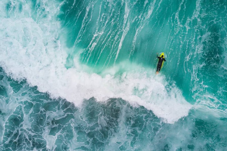 A surfer on a lime-green board rides beside a breaking wave in turquoise water, seen from above.