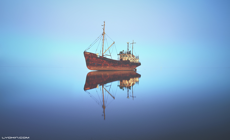 Rusting ship reflected in still blue water under a pale dawn sky.