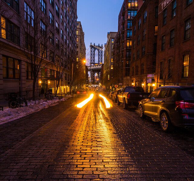 Car light trails on a cobblestone street leading toward the Manhattan Bridge at blue hour