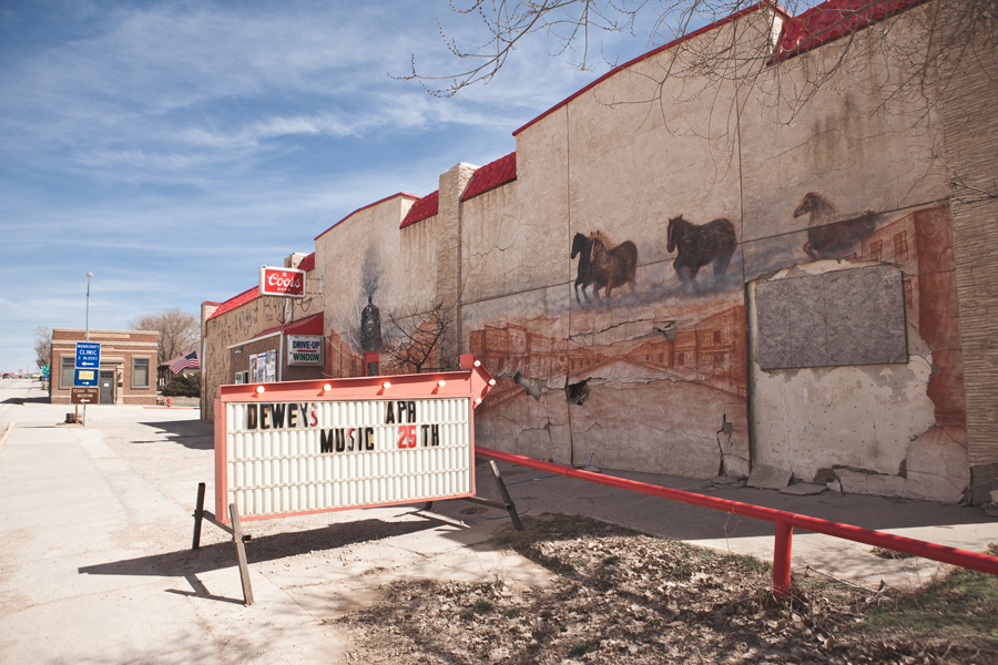 Weathered wall with a faded mural of running horses, beside a portable sign for Dewey's music and a Coors sign.