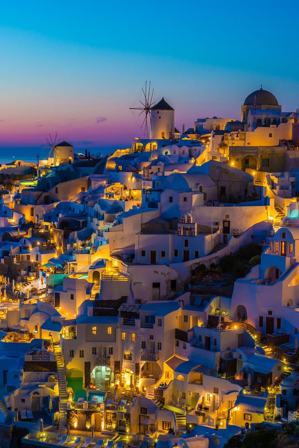 Windmills and illuminated white buildings in Oia, Santorini, at blue hour.