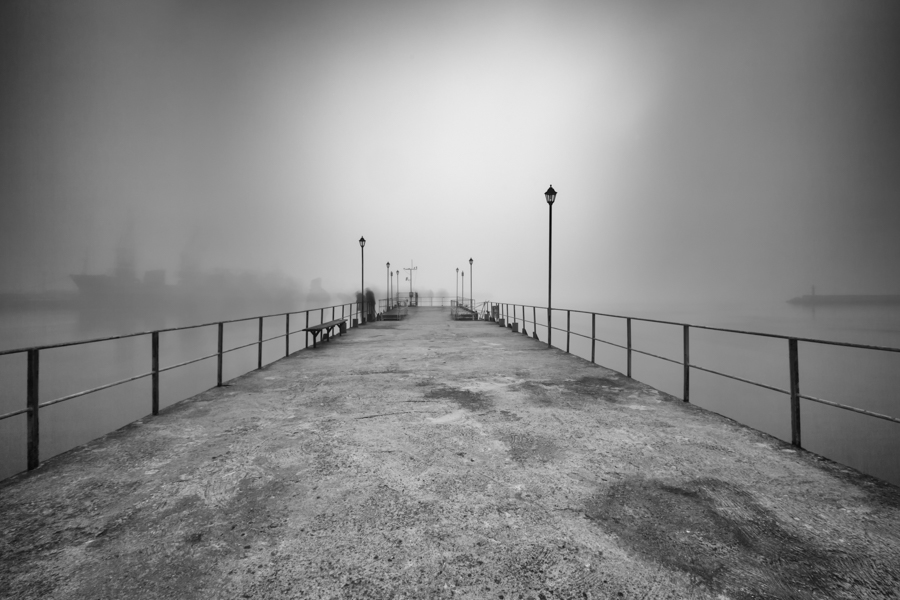 Empty stone pier disappearing into fog with railings on both sides.
