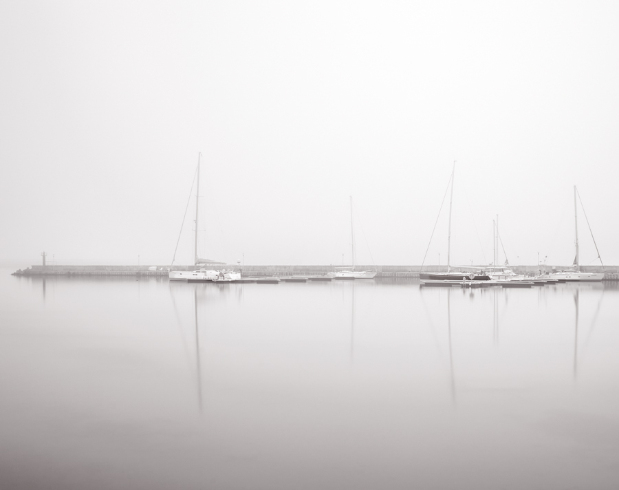 Sailboats reflected in glassy water beneath dense fog.
