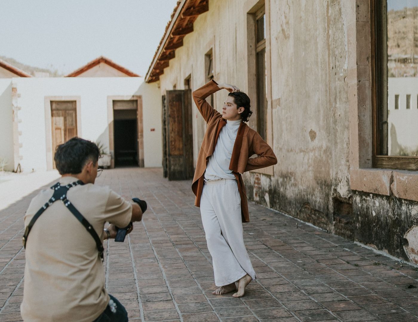 Photographer kneels with a camera facing a woman posing with one arm raised in an outdoor courtyard.