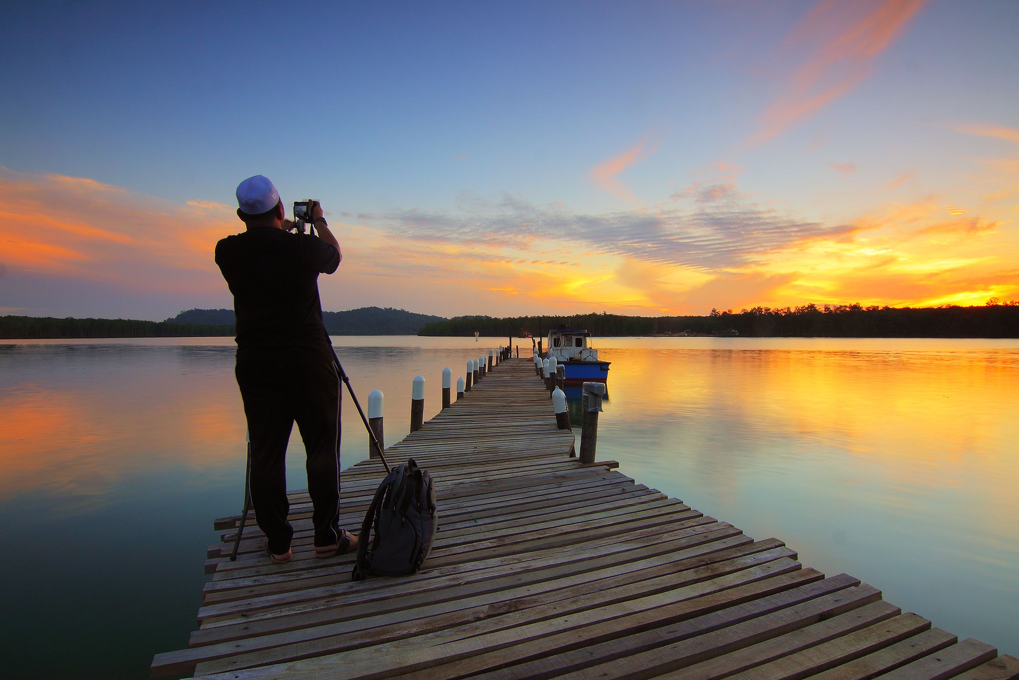 Photographer with a tripod and a backpack standing on a wooden pier over a lake at sunset, photographing the colorful sky