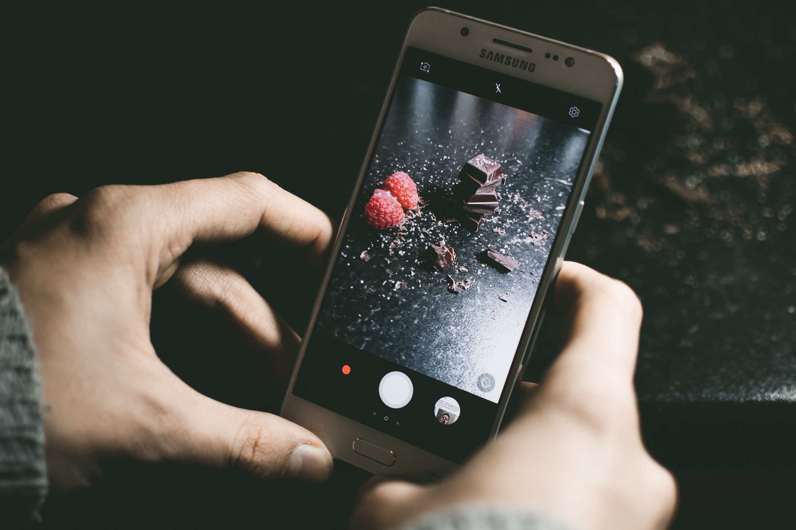 Hands hold a phone camera over berries and chocolate arranged on a dark tabletop