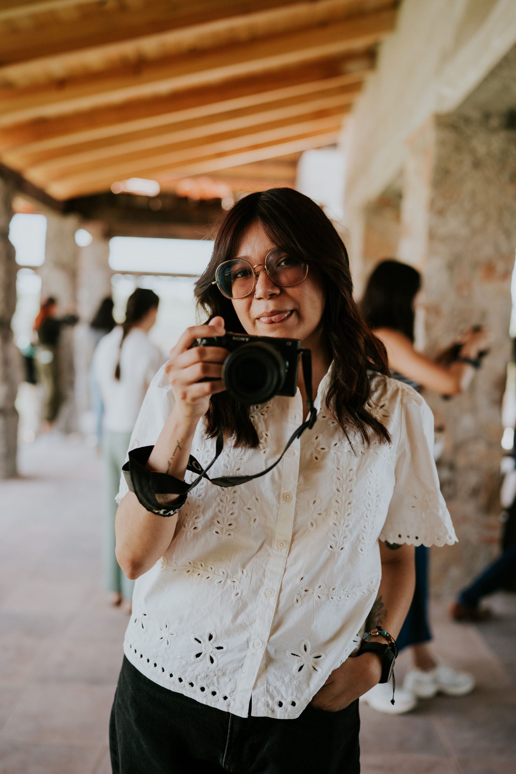 A woman in a white blouse holding a camera among guests at an indoor event.