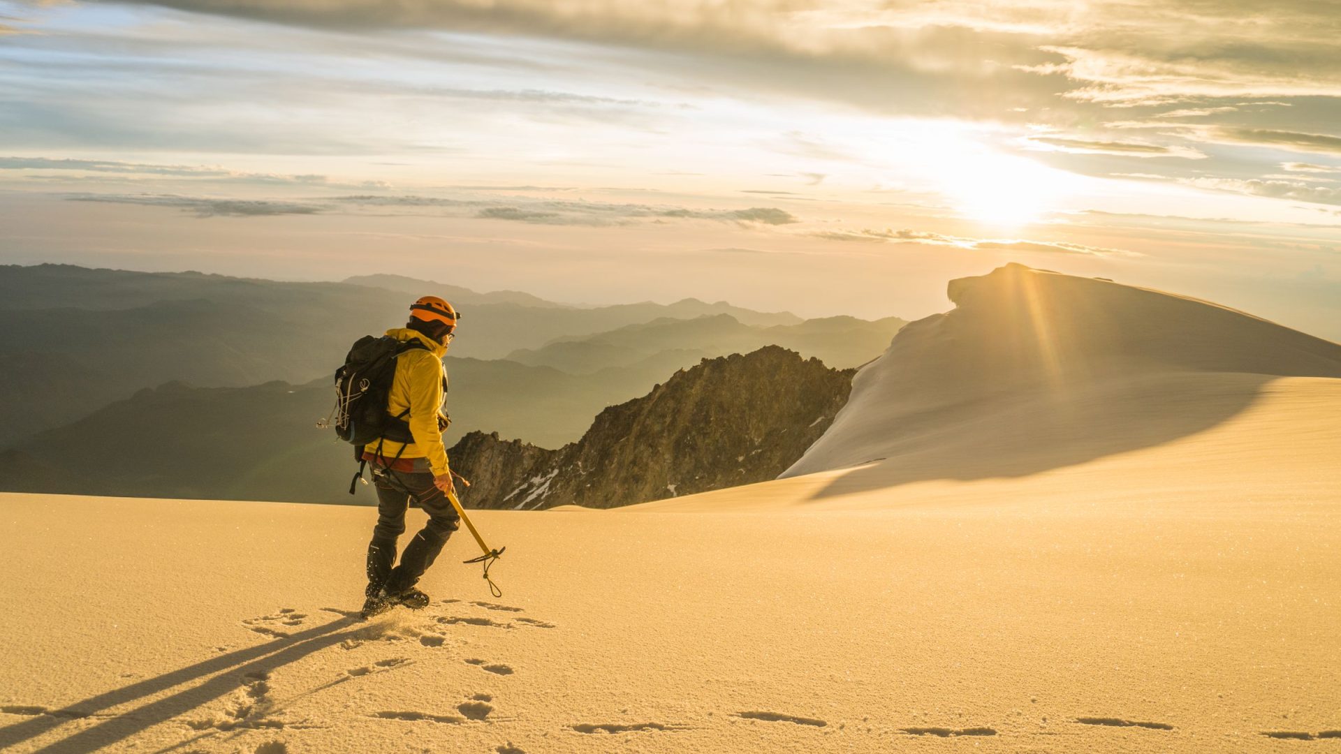 Mountaineer in a yellow jacket and backpack crosses a snowy ridge at golden hour, with mountains in the distance.