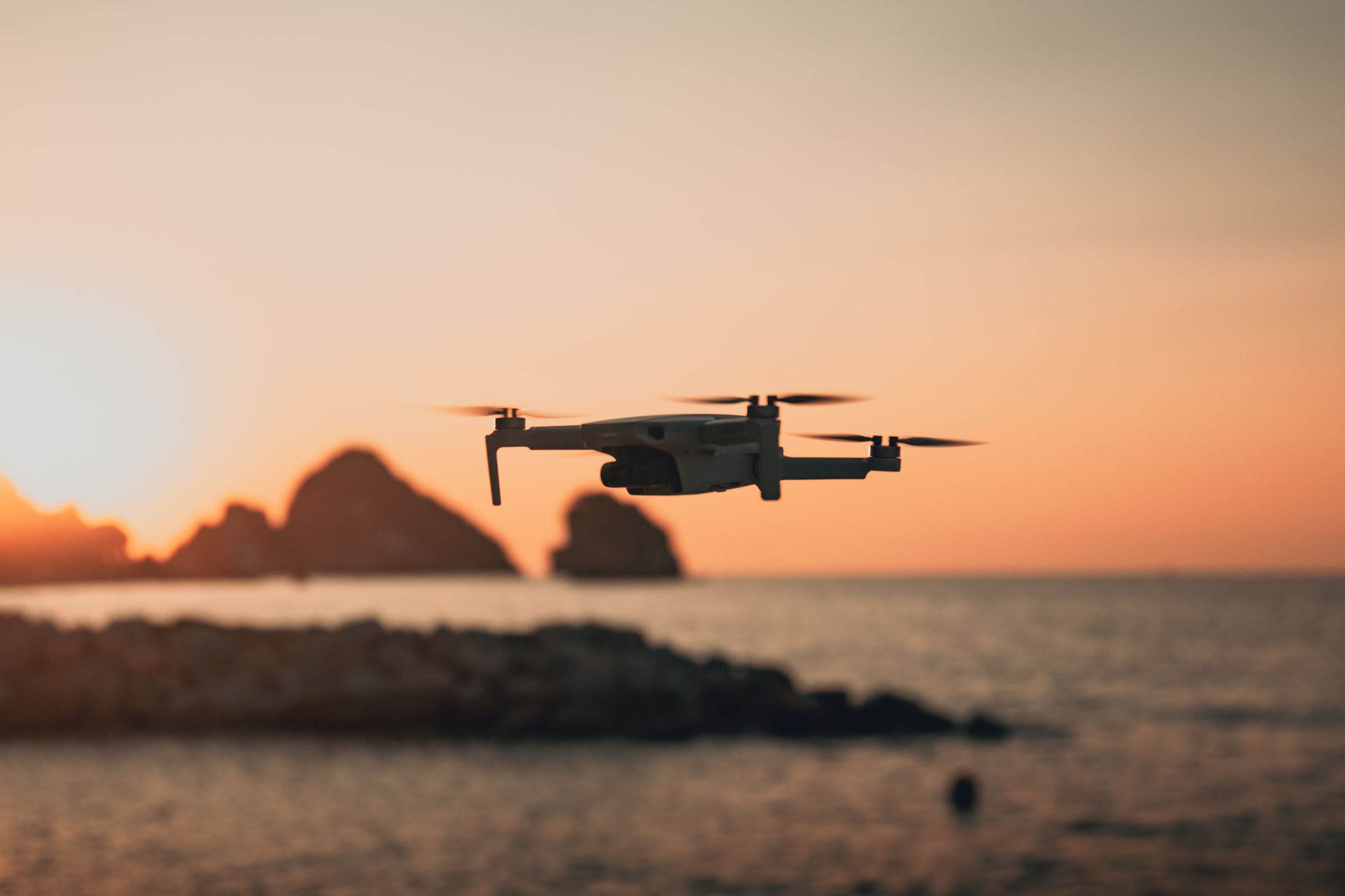 Quadcopter drone flying above a coastline at sunset, with rock formations offshore.