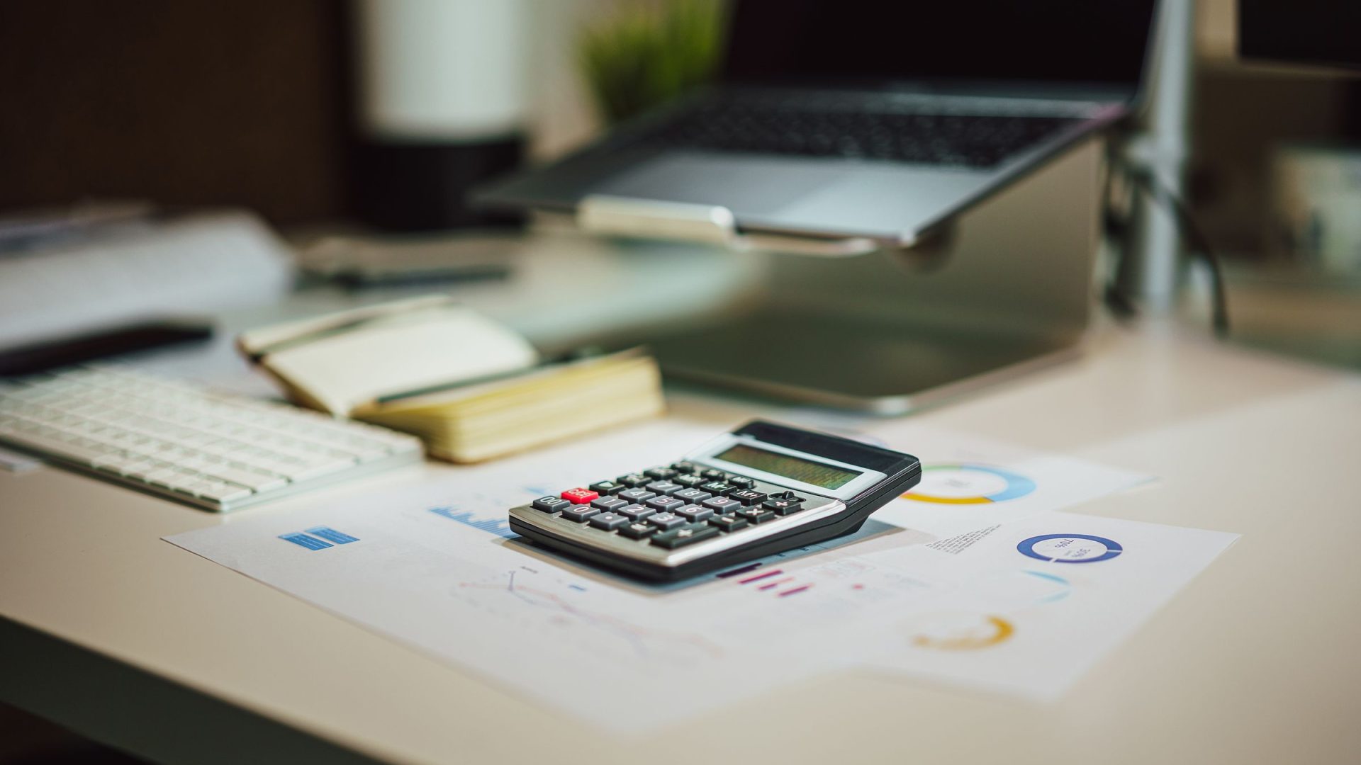 Calculator on financial charts beside a laptop and notebook on a work desk