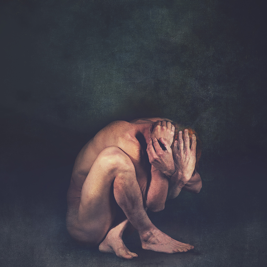 A nude figure crouches against a dark background with their face covered.