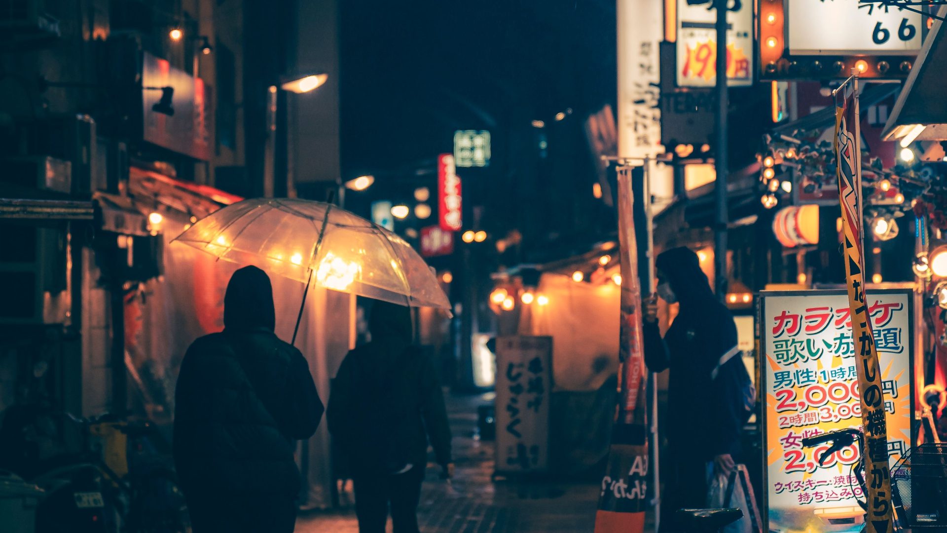 Umbrella-carrying pedestrians on a wet city street under glowing signs at night