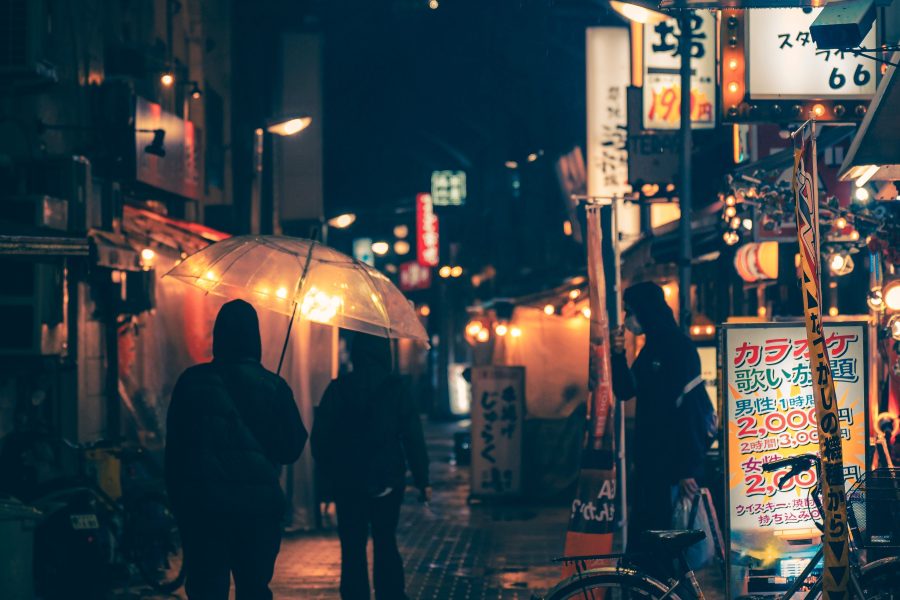 Umbrella-carrying pedestrians on a wet city street under glowing signs at night