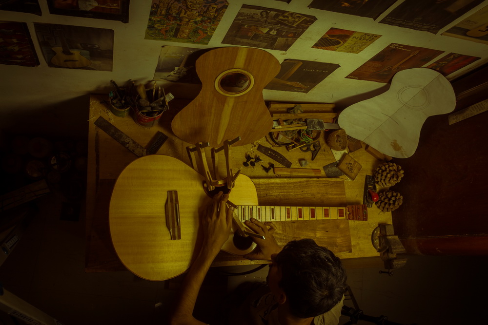 Overhead view of a guitar maker working at a crowded workshop table.