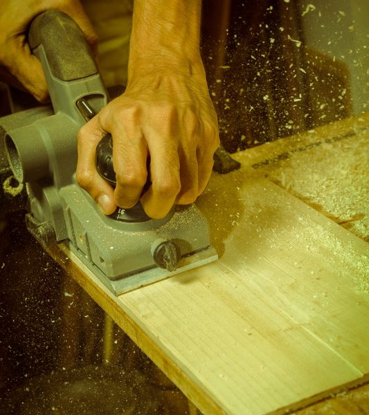 Hands guide a power tool along a wooden board, scattering sawdust.