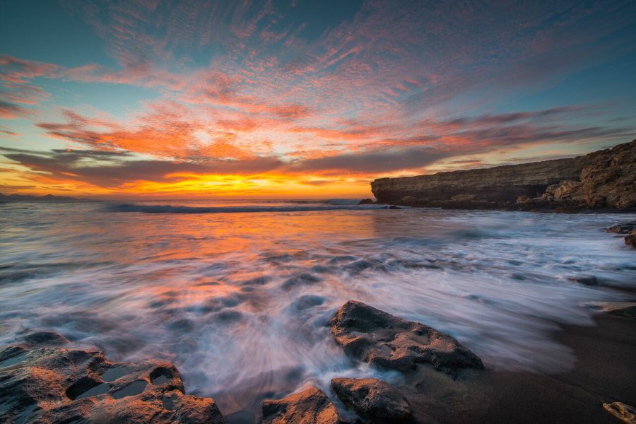 Long-exposure surf washing around rocks beneath a golden and pink sunset sky.