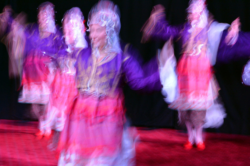Folk dancers in bright costumes move across a red stage in a long exposure.
