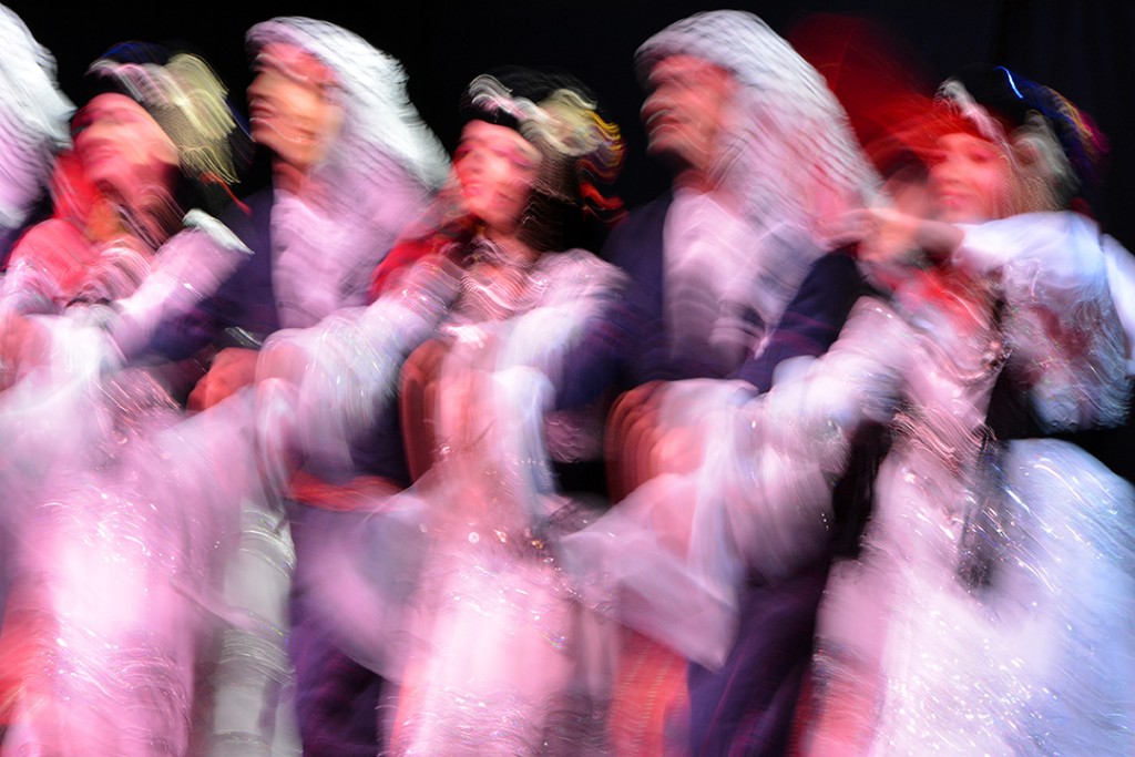 Performers in pink and white costumes move across a dark stage, blurred by a long exposure.