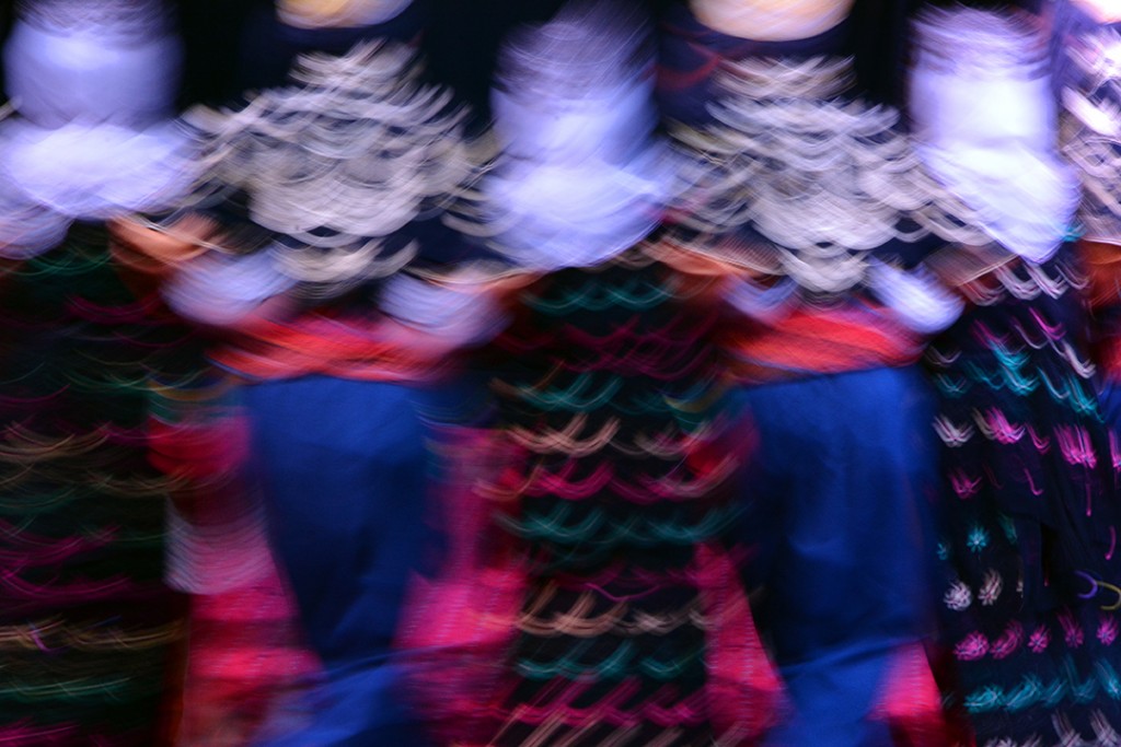 Dancers in white embroidered tops and dark skirts create streaks of motion against a black background.