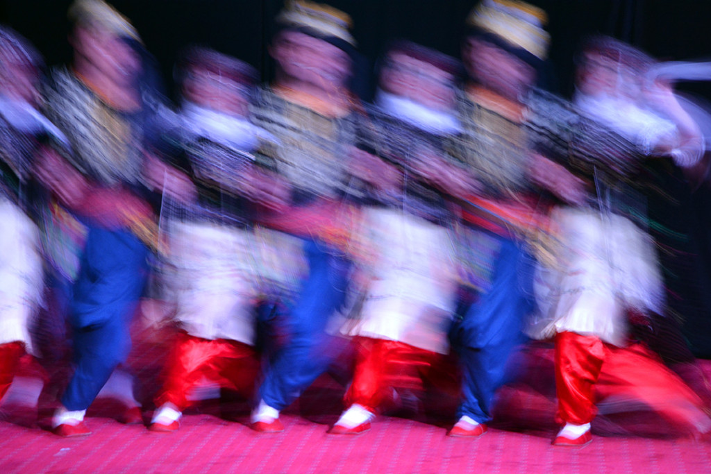 Row of dancers in embroidered costumes and bright red pants blurred in motion.