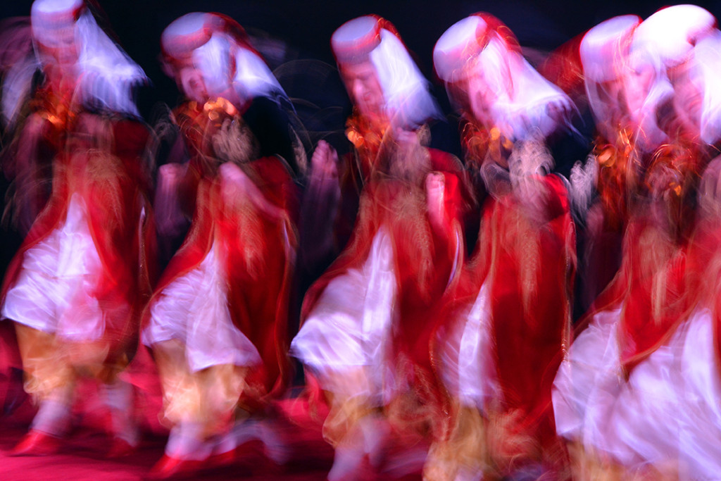 Dancers in red dresses and white head coverings appear as overlapping shapes in a long exposure.