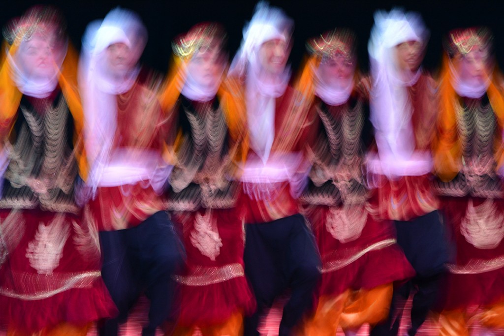 Folk dancers in orange, red, and white costumes moving across a dark stage.