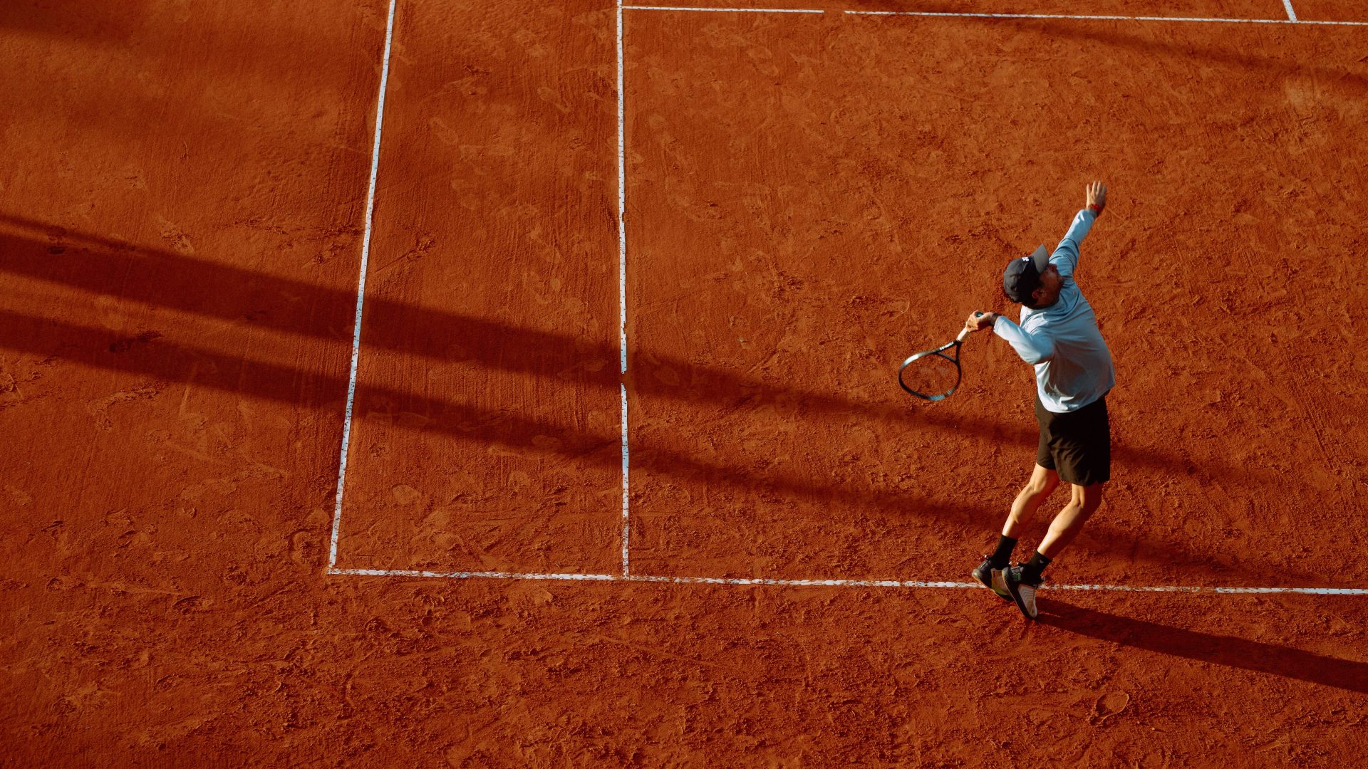 Tennis player serving on a red clay court in low sunlight