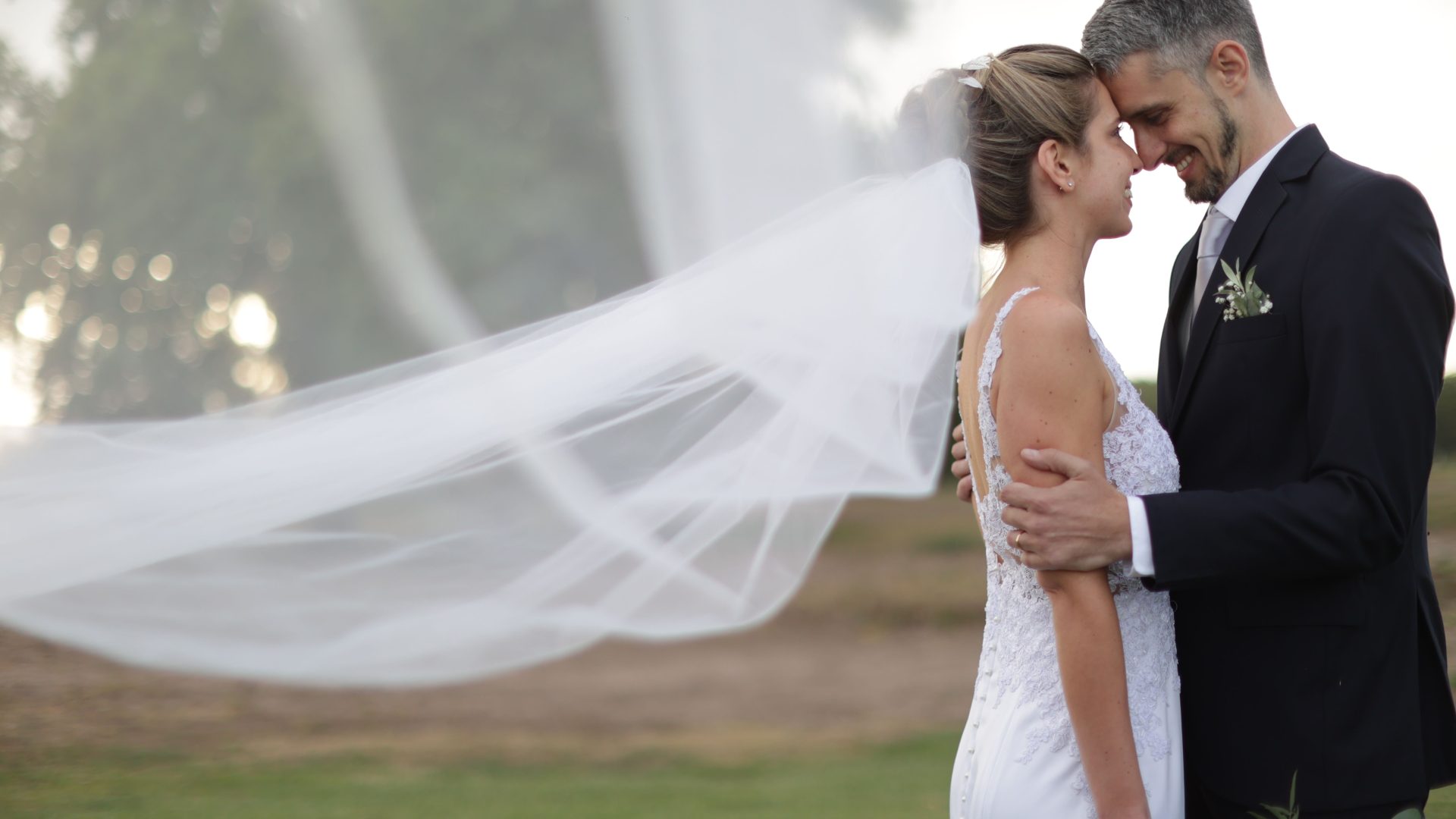 Bride and groom touching foreheads outdoors, with her veil flowing behind them