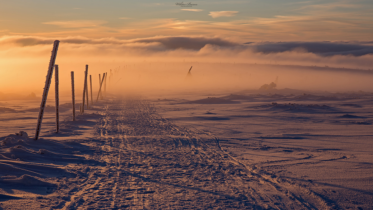 Wooden fence posts crossing a snow-covered mountain ridge at sunset.
